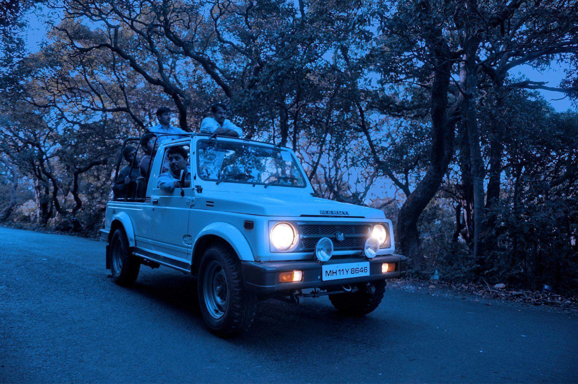 A white open-top Maruti Gypsy jeep with passengers is driving on a forested road near Ramsukh Resort in Mahabaleshwar. The vehicle is illuminated under a blue evening sky, with tall trees and dense foliage lining both sides of the road, creating a peaceful, adventurous atmosphere. The headlights of the jeep are on, and the license plate is visible with the number MH11Y 0848